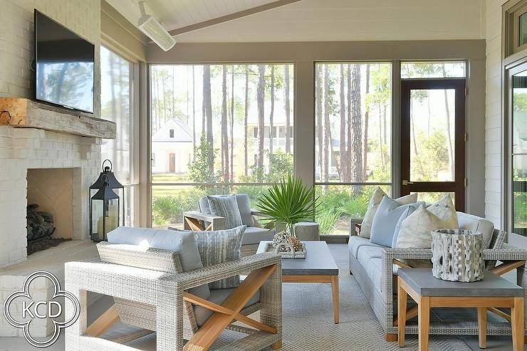 Welcoming gray sunroom features a gray wicker and teak couch placed on a gray rug beside a teak and concrete end table and facing a teak and concrete coffee table. The coffee table is flanked by gray wicker and teak accent chairs, while the space is completed with a white painted brick fireplace finished with a rustic wood mantel and wall mounted TV.