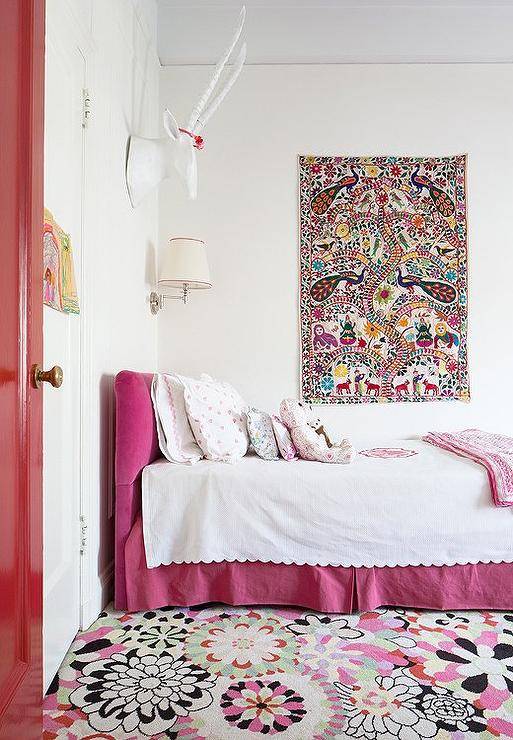 Pink girl's bedroom features a faux deer head doubling as a headband holder situated over a pink velvet headboard on twin bed dressed in white scalloped with pink monogram blanket, pink heart shams and a white and pink blanket placed at the foot of the bed as well as a pink pleated bedskirt situated under a colorful tapestry and atop floral carpeting.