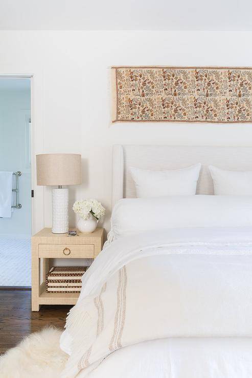 This gorgeous bedroom features a restful ambiance starting with a white wingback headboard dressed in white and tan linen bedding. A gold grasscloth wrapped nightstand displays a white greek key lamp with surrounding accents from a long tapestry over headboard and white faux fur rug.