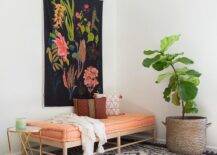 A botanical tapestry hangs over a wood daybed accented with a sherbet tufted cushion and placed on a black stripe fringe rug between a large potted plant and a black and brass accent table.