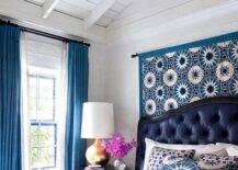 White plank vaulted ceiling holds a glass and brass Moravian star pendant over a bed dressed in a yellow medallion blanket accented with white sheets topped with a blue medallion pillow layered in front of blue and red print pillows and matching a tapestry hung behind the navy blue leather tufted headboard. Beside the bed, a round black beside table lit by a gold leaf table lamp positioned beside a window dressed in ocean blue linen curtains.