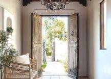 Mediterranean style foyer features a turquoise blue overdyed rug on a terracotta grid floor with a rattan bench and a reclaimed wood double front doors illuminated by a wrought iron chandelier hung from a ceiling with ornate ceiling beams.