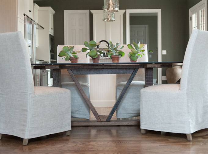 Hannah Maple - Beautiful dining room with dark taupe walls paired with crisp white trim. The dining room features a trestle style dining table with glass top. The dining table is lined with linen slipcovered dining chairs. Four succulents in terracotta pots stand upon the dining table. A pair of nickel lantern pendants hang over the dining table. Hardwood floors complete the space.