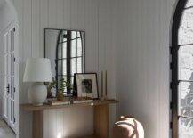 Foyer features a tan console table illuminated by an Aerin Toulon table lamp, a black mirror leaning on vertical plank walls and a tall terracotta vase on the floor.