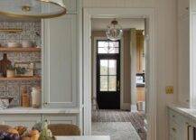 Red brick herringbone pavers lead to a black mudroom door finished with glass panels and a brass door handle and located beneath a transom window.