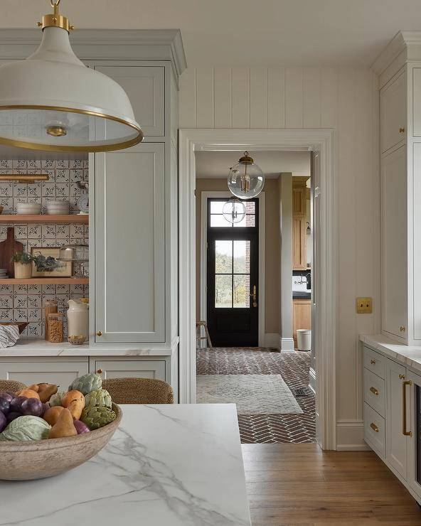 Red brick herringbone pavers lead to a black mudroom door finished with glass panels and a brass door handle and located beneath a transom window.