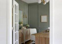 White French doors under an arched transom window opens to a beautifully styled brown and green home office featuring a brown wooden desk placed on a white and brown rug with a white task chair. The desk is lit by a white and gold sputnik light while green walls are accented with green trim moldings.