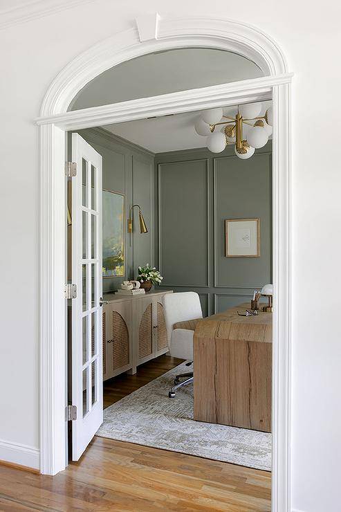 White French doors under an arched transom window opens to a beautifully styled brown and green home office featuring a brown wooden desk placed on a white and brown rug with a white task chair. The desk is lit by a white and gold sputnik light while green walls are accented with green trim moldings.