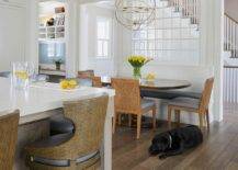 A sphere chandelier lights an oval pedestal dining table matched with fabric and abaca dining chairs and a white built-in dining bench with a black leather cushion. An indoor window is located over the bench and beneath a transom window.