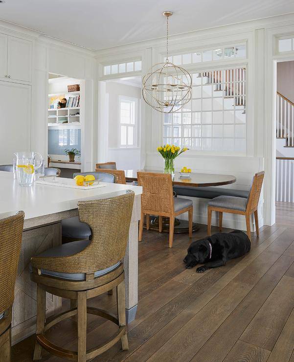 A sphere chandelier lights an oval pedestal dining table matched with fabric and abaca dining chairs and a white built-in dining bench with a black leather cushion. An indoor window is located over the bench and beneath a transom window.