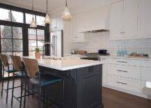 Two-tone vintage island stools at a black center island under antiqued mercury glass lights pendants. White quartz countertops balance the black island while the perimiter space is equipped with horizontal white tiles, a white vent hood, and chrome hardware.