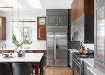 Kitchen features a brown reeded and gray marble hood over a cooktop and gray velvet dining chairs at a brown wooden center island with sink that boasts a brass gooseneck faucet illuminated by staggered linear island lights and gray and brown two tone kitchen cabinetry.