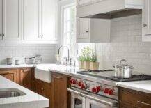 White upper cabinets with brown lower cabinets brings a stunning two-toned appeal to a transitional kitchen. A white wood panel range hood mounts above white subway tiles and a wolf dual range. A sink next to the stove features a polished nickel gooseneck faucet with a window overhead bringing nautral light to the kitchen design.