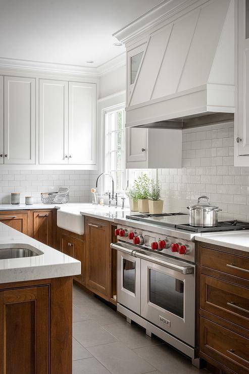 White upper cabinets with brown lower cabinets brings a stunning two-toned appeal to a transitional kitchen. A white wood panel range hood mounts above white subway tiles and a wolf dual range. A sink next to the stove features a polished nickel gooseneck faucet with a window overhead bringing nautral light to the kitchen design.