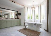 Stunning bathroom features an extra-long green washstand topped with gray and white marble placed under a gray wash mirror lined with Ralph Lauren Singleton Sconces. A marble herringbone tiled floor leads to bow window dressed in white and taupe curtains filled with a roll top bathtub and a faux bois accent table illuminated by a Restoration Hardware Victorian Hotel Pendant.