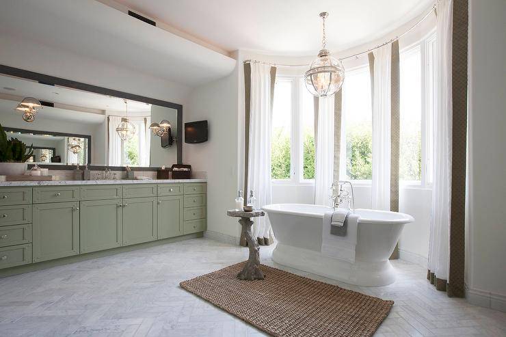 Stunning bathroom features an extra-long green washstand topped with gray and white marble placed under a gray wash mirror lined with Ralph Lauren Singleton Sconces. A marble herringbone tiled floor leads to bow window dressed in white and taupe curtains filled with a roll top bathtub and a faux bois accent table illuminated by a Restoration Hardware Victorian Hotel Pendant.