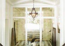 Stunning black and white foyer is illuminated by a Restoration hardware Victorian Hotel Pendant hung from a white ceiling accented with an x-trim over a round ivory leather ottoman placed on white and black harlequin floor tiles in front of a mirrored paneled wall.