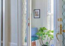 Leaded sidelights and a leaded glass transom window frame a white door open to a long and narrow vestibule boasting a white built-in bench topped with a turquoise blue cushion accented with a white and blue pillow placed against a shiplap wall.