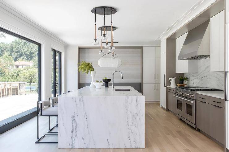 An eye-catching staggered chandelier is mounted over a white and gray marble waterfall edge kitchen island seating black and white stools.