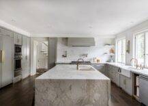 A sliding cutting board accents a kitchen sink with brass gooseneck faucets mounted to a marble waterfall edge island.