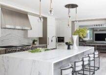 A row of black and white stools sit at a white and gray marble waterfall edge island lit by staggered chandeliers.