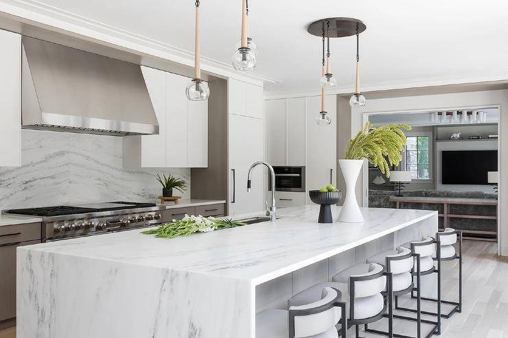 A row of black and white stools sit at a white and gray marble waterfall edge island lit by staggered chandeliers.