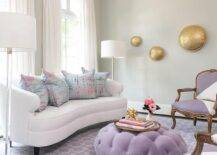 Arched windows framed by a light gray wall and covered in white curtains are positioned behind a white curved sofa topped with pink and blue pillows and placed on a purple chain link rug between glass and nickel floor lamps. White and purple French chairs sit facing a purple velvet tufted ottoman coffee table placed in front of the sofa.