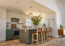 White and gray kitchen features white and gold globe pendants that illuminate a dark gray center island with brown woven leather stools, white cabinets with brass pulls and a white refrigerator.