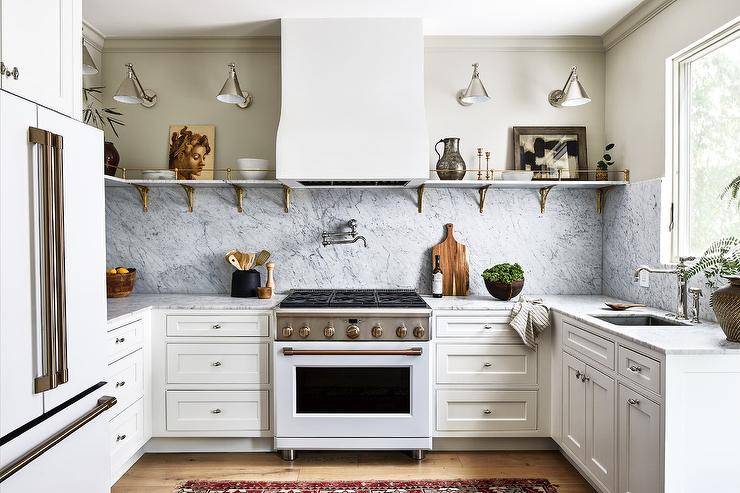 White and gray kitchen features long marble and brass shelves lit by a Boston functional library lights, a white hood over a nickel swing arm pot filler and white stove and marble backsplash, a white French door refrigerator and white cabinets.