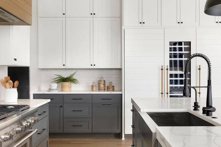 Well-appointed white and gray kitchen features dark gray lower cabinets accented with oil rubbed bronze pulls and a marble-like countertop fixed against a white exposed brick backsplash and beneath stacked white shaker upper cabinets finished with orb knobs. A hidden white shiplap fridge with a glass front door is inset beneath white shaker cabinets.