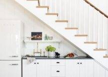 Beneath a two-toned staircase, white plank kitchenette cabinets are accented with oil rubbed bronze hardware and a black marble countertop fitted with a sink and a polished nickel deck mount faucet kit. A small white shelf is mounted to white shiplap trim over the sink and beside a white Smeg fridge.