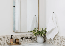 A powder room wall half clad in white and brown mosaic tiles features a brown single washstand donning brass knobs and a marble countertop holding a sink beneath a wall mount oil rubbed bronze vintage faucet. The faucet is fixed beneath a curved brass mirror illuminated by a vintage brass sconce.