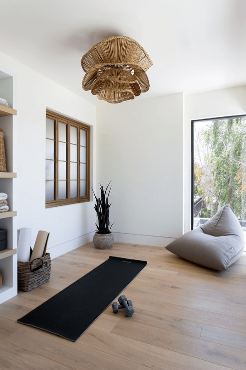 A stunning wicker chandelier is fixed over a black yoga mat placed on a wood floor, while a gray floor pillow is placed in front of a picture window.