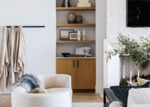 Living room features a white curved sofa accented with a beige velvet tufted ball pillow, a wall mount throw blanket rack and a built in shelf next to a fireplace.