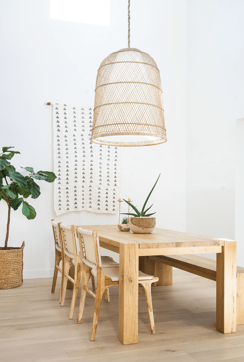 A large basket chandelier lights a chunky oak dining table paired with white leather and wooden dining chairs and positioned in front of a white and gray geometric tapestry hung from a white wall.