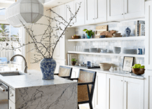 A white and gold linear light hangs from a blue tray ceiling over a black and white marble waterfall edge island paired with cane stools.