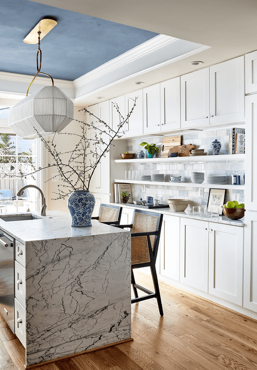 A white and gold linear light hangs from a blue tray ceiling over a black and white marble waterfall edge island paired with cane stools.