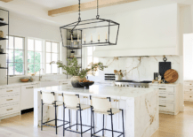 Darlana Linear Chandelier lights a white shaker island topped with a marble waterfall edge countertop seating ivory leather stools.