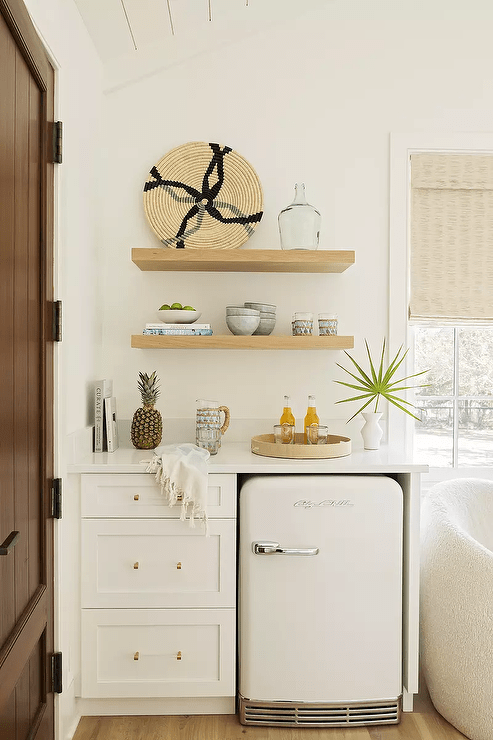 Stacked oak shelves are mounted over white kitchenette cabinets fitted in a bedroom and finished with a white mini Big Chill Fridge.